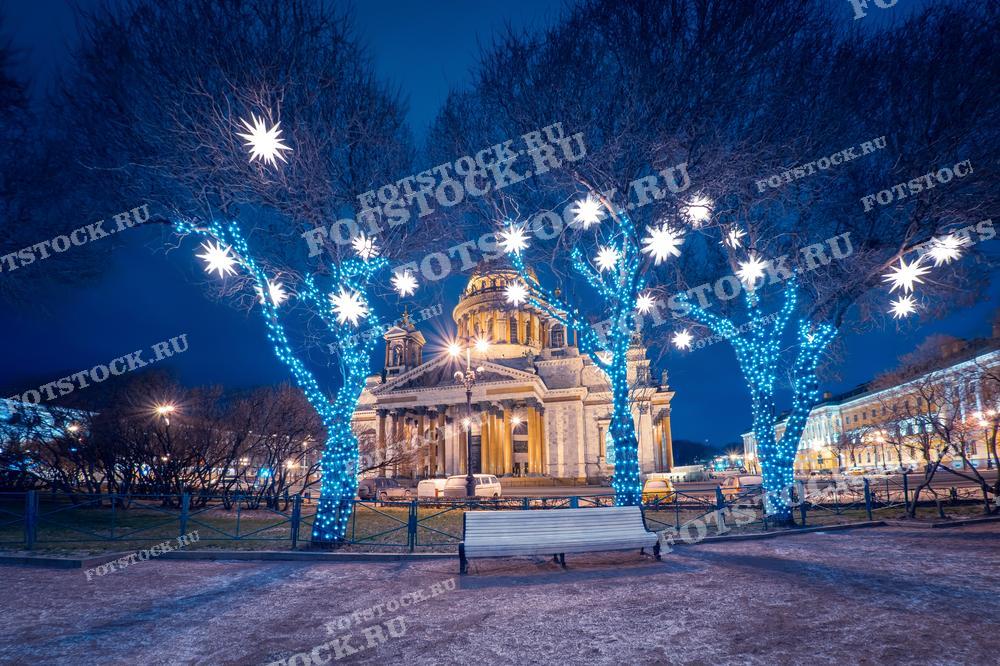 На фотографии изображен Исаакиевский собор в Санкт-Петербурге с новогодней иллюминацией на деревьях в вечернее время. Светящиеся украшения создают праздничную атмосферу. Фотография идеально подходит для использования в материалах о новогодних праздниках, туристических буклетах и статьях о зимнем Санкт-Петербурге.